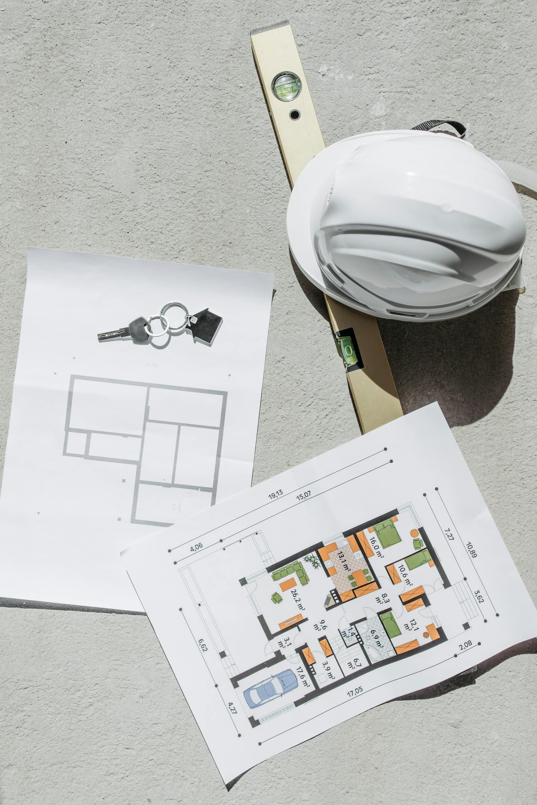 Flat lay of construction plans, keys, hard hat, and level tool on concrete.